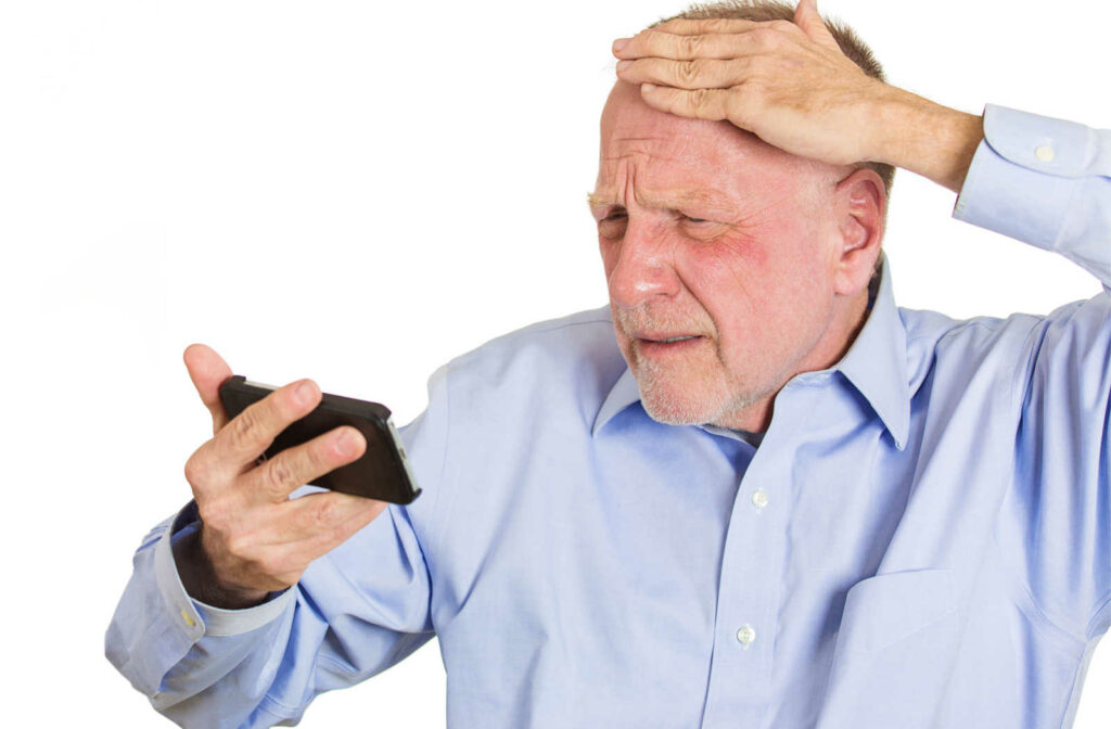 A close-up of a senior man holding his cell phone in his hand while squinting and touching his forehead, having difficulty seeing what is on the phone screen
