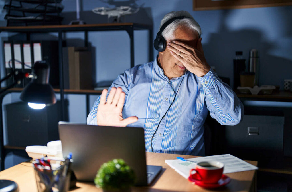 A grey-haired man is covering his eyes with his left hand, to shield light coming out of the computer screen.