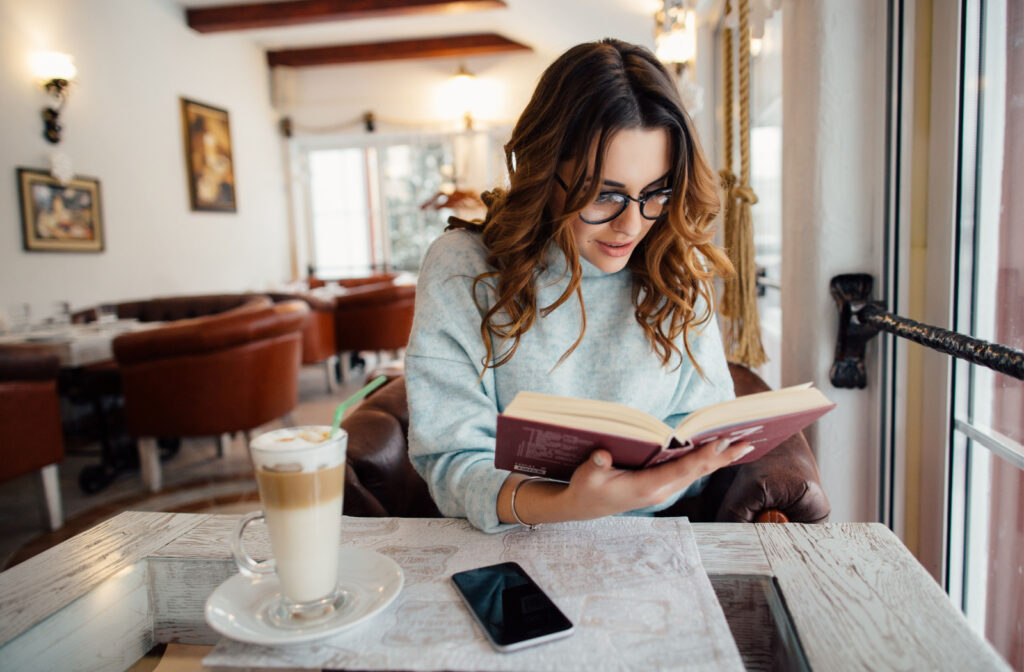 A woman with glasses reading a book while sitting alone in a beautiful restaurant. With a coffee and her cellphone on the table in front of her.